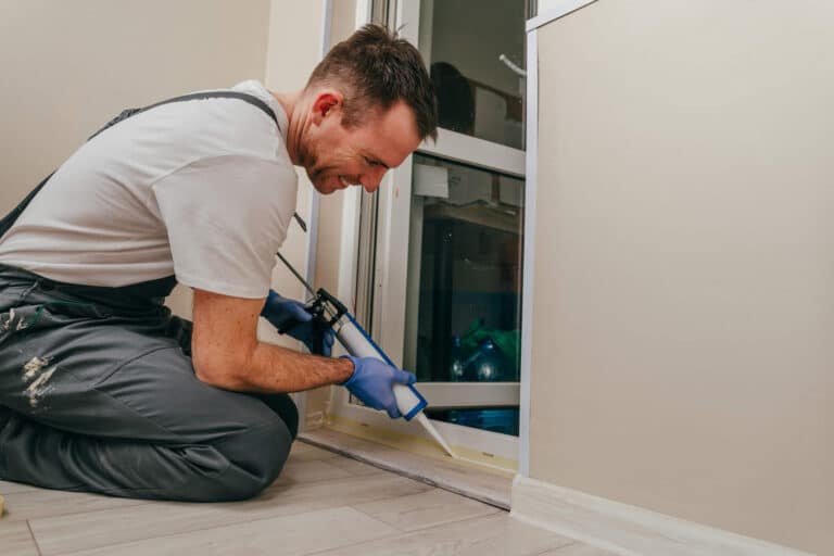 Young man wearing overalls sealing a door with caulk to prevent pests during the winter