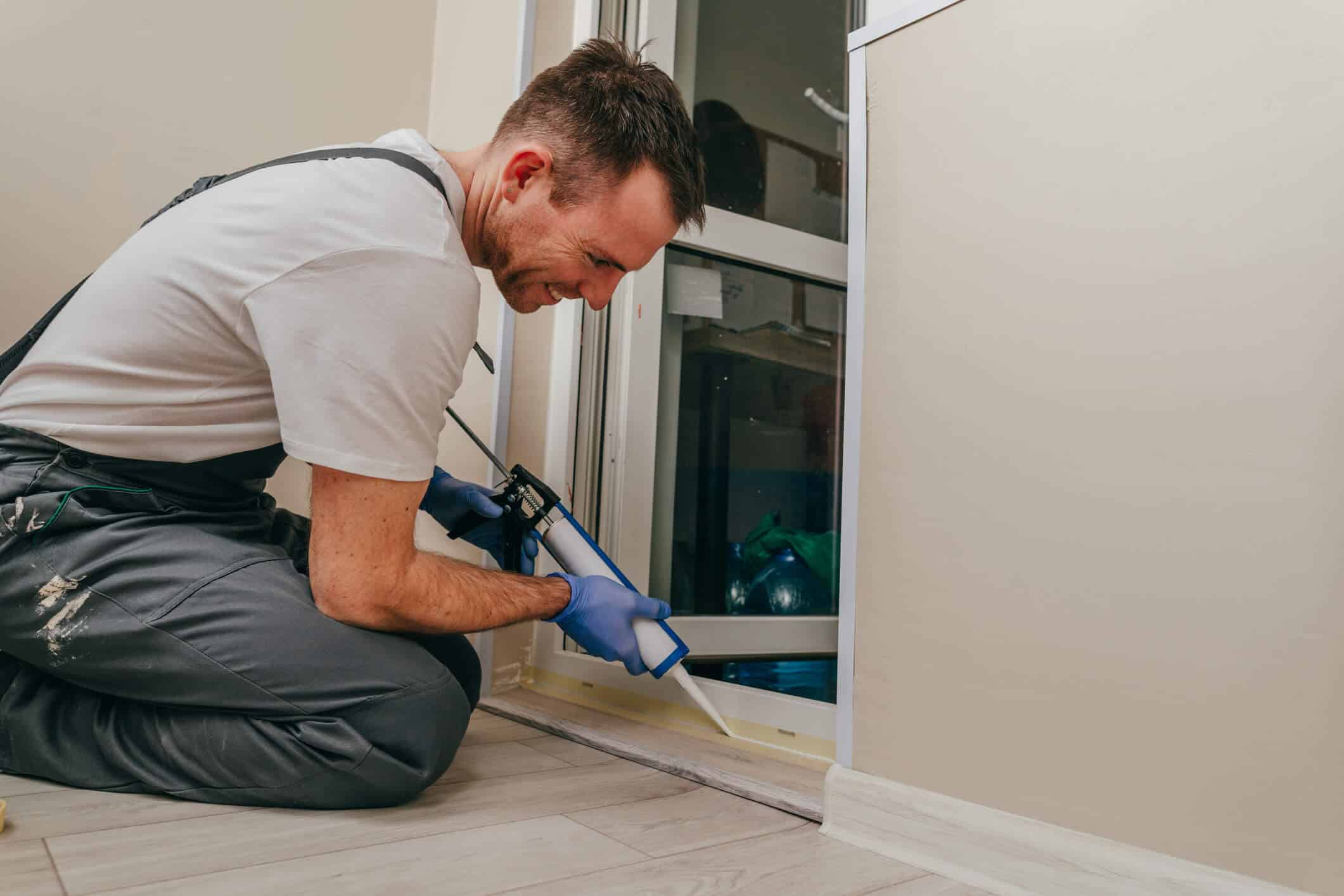 Young man wearing overalls sealing a door with caulk to prevent pests during the winter